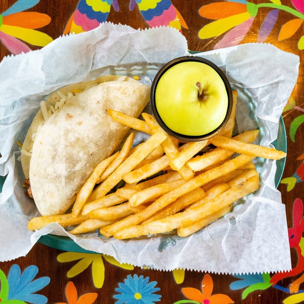 Taco, french fries, and a green apple served in a basket on a colorful table.