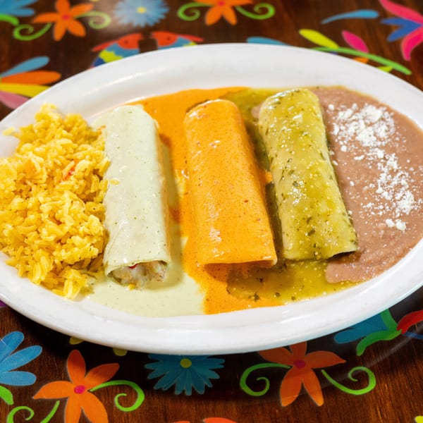 Three enchiladas with white, orange, and green sauces, rice, and beans on a colorful floral table.
