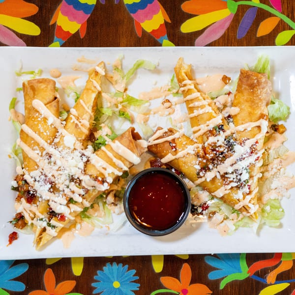Golden taquitos with lettuce, cotija cheese, and creamy sauce, served with a side of red dipping sauce.