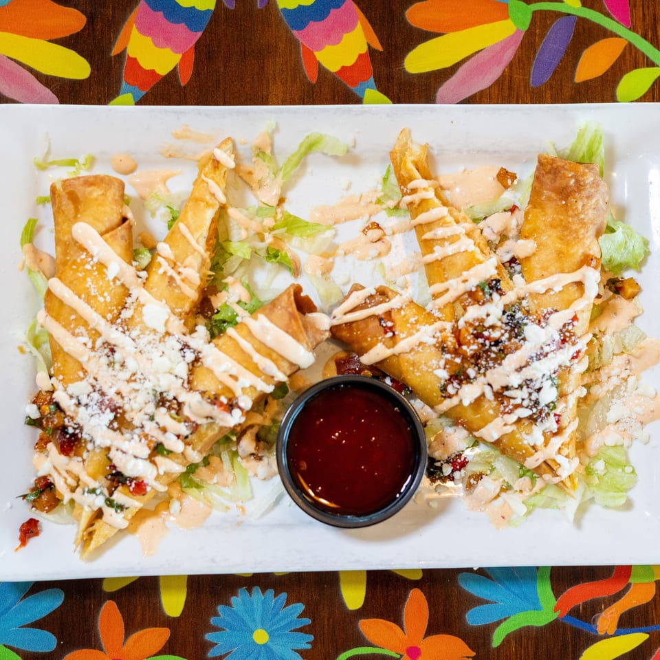 Golden taquitos with lettuce, cotija cheese, and creamy sauce, served with a side of red dipping sauce.