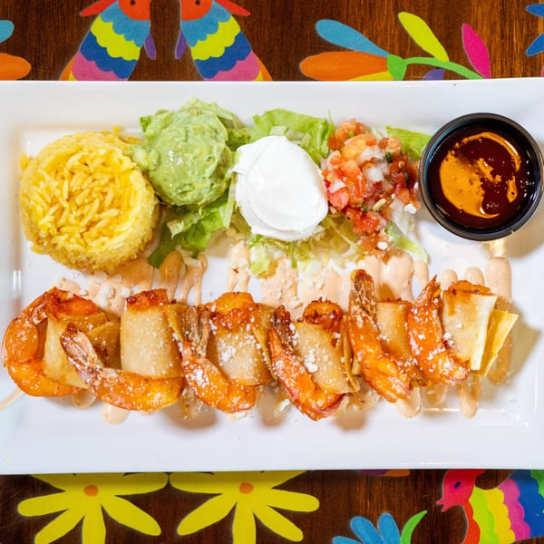Fried shrimp wrapped in pastry with yellow rice, guacamole, sour cream, pico de gallo, and sauce on a festive table.