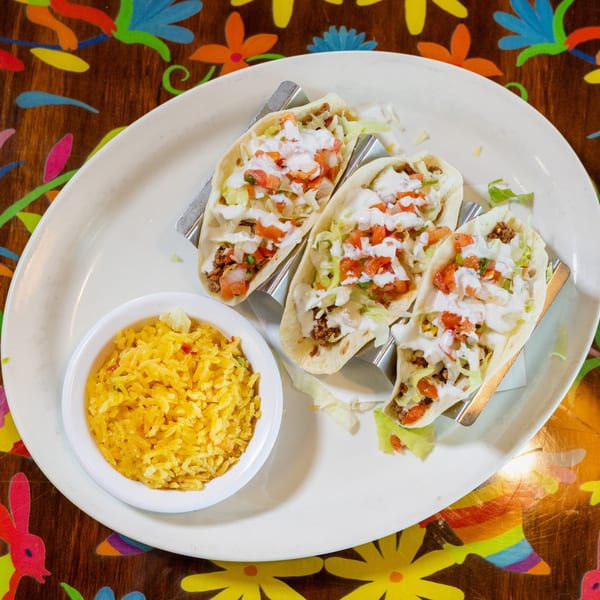 Three tacos with meat, lettuce, pico de gallo, and crema, served with yellow rice on a colorful table.