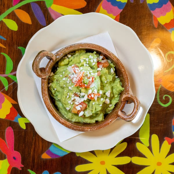 Guacamole in a rustic bowl with cheese and pico de gallo, on a white plate and colorful wooden table.