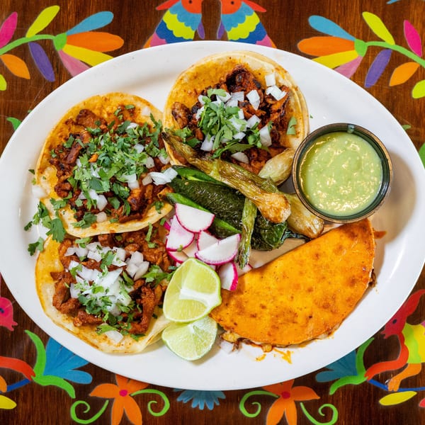 Plate of three tacos, a quesabirria, grilled peppers, radishes, lime, and green salsa on a colorful table.