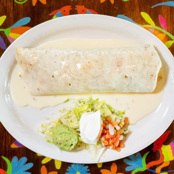 Burrito covered in white sauce with guacamole, sour cream, pico de gallo, and lettuce on a colorful table.