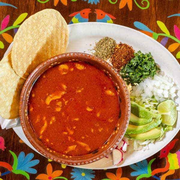 A vibrant bowl of red soup with fresh avocado, lime, onion, cilantro, and spices, alongside tostadas.