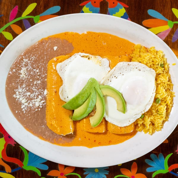 Enchiladas with orange sauce, fried eggs, avocado, refried beans, and yellow rice on a colorful table.