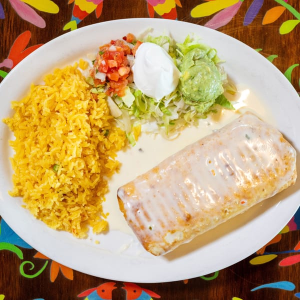 Burrito covered in white sauce, yellow rice, pico de gallo, sour cream, and guacamole on a white plate.