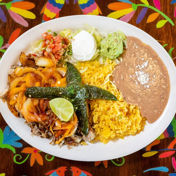 Vibrant Mexican combo plate with sautéed meat, onions, rice, beans, guacamole, and chiles on a patterned table.