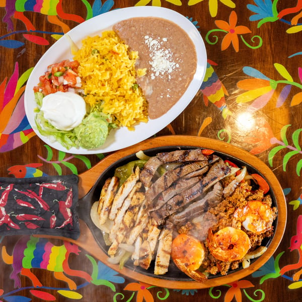 Sizzling fajita platter with steak, chicken, shrimp, and a side of rice and beans on a colorful table.